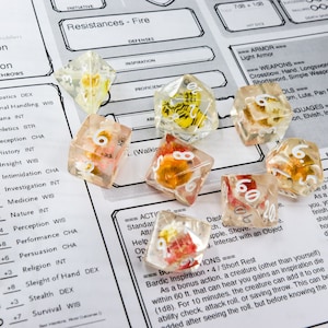 Puede incluir: Un conjunto de dados de resina transparente con inclusiones coloridas, incluyendo flores. Los dados están dispersos sobre una hoja de juego impresa, ideal para juegos de rol.
