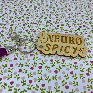 Puede incluir: Llavero de madera con un borla morado. El llavero tiene el texto "NEURO SPICY" en &eacute;l.