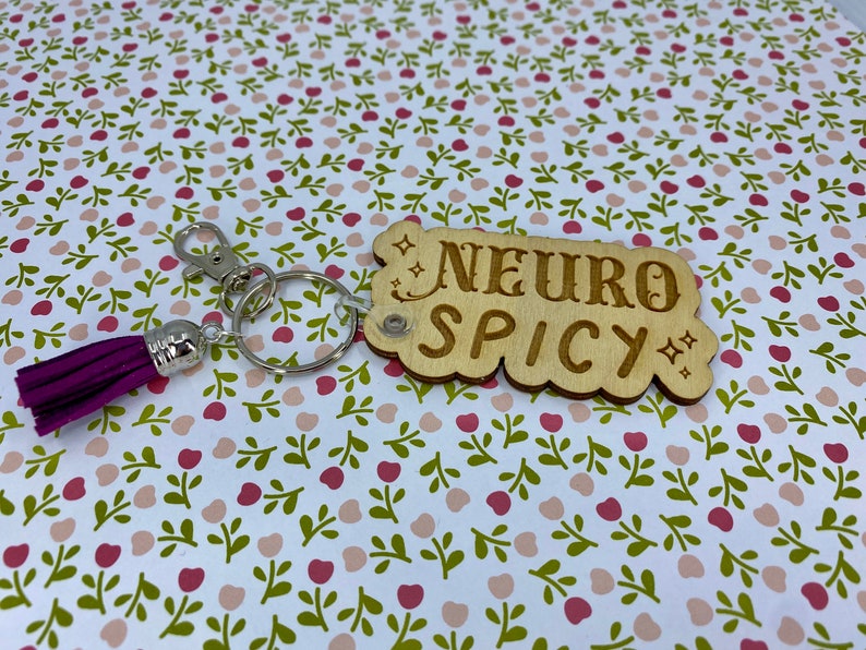 Puede incluir: Llavero de madera con un borla morado. El llavero tiene el texto "NEURO SPICY" en &eacute;l.