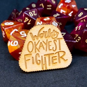 Puede incluir: Un token de madera con borde festoneado con la inscripción "World's Okayest Fighter" se encuentra frente a una colección de dados poliédricos. Los dados son de varios tamaños y colores, incluyendo rojo, naranja y dorado.