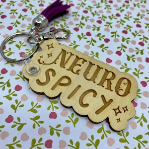 Puede incluir: Llavero de madera con un borla morado y el texto "Neuro Spicy" en una fuente vintage.
