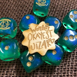 May include: A collection of translucent teal and blue dice with gold-colored pips and symbols. A wooden star-shaped piece in the center reads "World's Okayest Wizard". The dice are arranged on a textured brown surface.