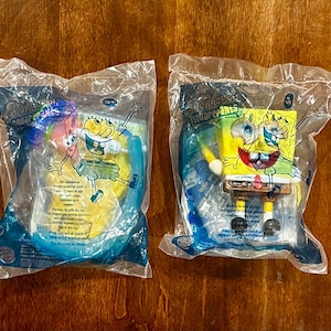 Op de afbeelding: Twee verzegelde plastic zakjes met SpongeBob SquarePants speelgoed. Het linkerzakje bevat een gele SpongeBob-figuur met een blauwe snorkel en duikbril. Het rechterzakje bevat een gele SpongeBob-figuur met een gele spatel en bruine broek.