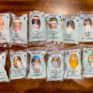 May include: A selection of Alexander doll toys from McDonald's Happy Meals, each in a clear plastic bag. The packaging has a light blue design and the McDonald's logo. The dolls are in various outfits.