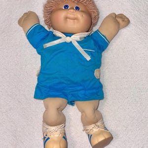 May include: A vintage Cabbage Patch Kid doll with a light skin tone, blue eyes, and a smiling expression. The doll wears a blue romper with white trim and a white bow. Its light brown yarn hair is styled in a short, curly fashion. The doll is wearing beige shoes with blue stripes.