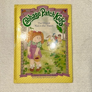 May include: A vintage Cabbage Patch Kids storybook. The cover features a girl in a purple dress and green top, holding a basket. The title reads "The Shyest Kid in the Patch." The book is framed in yellow with a purple banner.