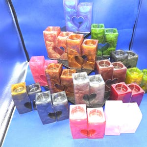 Könnte beinhalten: Eine Sammlung rechteckiger Kerzenhalter aus Kunstharz in verschiedenen Farben wie Blau, Grün, Rot, Pink, Orange und Grau. Jeder Halter hat eine herzförmige Aussparung auf der Vorderseite und eine kreisförmige Öffnung oben für eine Kerze.