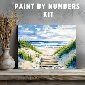 Puede incluir: Kit de pintura por números que presenta una escena de playa con una pasarela de madera que conduce al océano. El cielo es azul con nubes blancas, y la arena es blanca. El océano es azul con olas blancas.