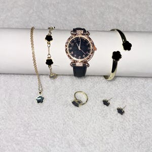 Puede incluir: Un conjunto de joyas doradas y un reloj con detalles en negro. El conjunto incluye un collar, una pulsera, un anillo, pendientes, un reloj y una pulsera de puño, todos con un diseño de trébol. El reloj tiene una correa de cuero negro.