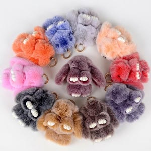 May include: A collection of eleven fluffy, plush rabbit-shaped keychains arranged in a circle. Each keychain features soft, faux fur in various colors including orange, blue, gray, peach, pink, red, purple, brown, and charcoal, with white feet.