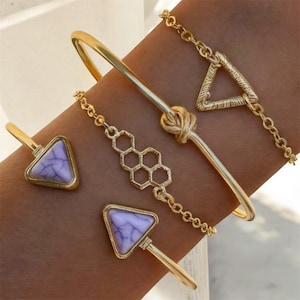 Puede incluir: Una colección de pulseras doradas. Una es una pulsera rígida con un diseño de nudo, y otras presentan formas geométricas como triángulos y un patrón de panal. Dos pulseras tienen detalles triangulares de piedra púrpura.