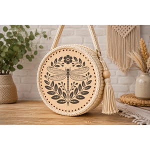 May include: Round, cream-colored handbag with a dragonfly design. The bag features a light-colored, circular leather panel with a black dragonfly and floral illustration. The bag has a woven texture, a shoulder strap, and a tassel.