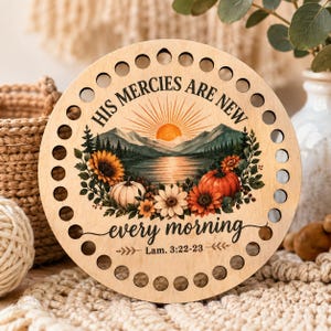 May include: A circular wooden plaque with a series of holes along the perimeter. The center features a printed landscape of mountains, a lake, and a sunrise, surrounded by sunflowers and pumpkins. Text reads: HIS MERCIES ARE NEW every morning Lam. 3:22-23.