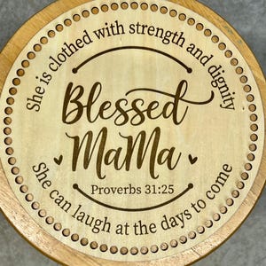 May include: A wooden round stool with a laser-engraved design. The design features the text "She is clothed with strength and dignity Blessed Mama She can laugh at the days to come Proverbs 31:25".