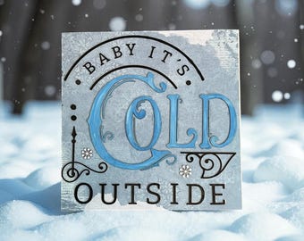 Può includere: Un cartello quadrato con la scritta "BABY IT'S COLD OUTSIDE" in lettere nere e blu. Il cartello ha uno sfondo azzurro e grigio con decorazioni a fiocco di neve ed è ambientato in un ambiente innevato.