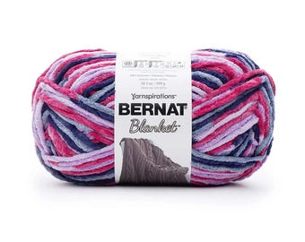Puede incluir: Una gran madeja de hilo Bernat Blanket en tonos rosa, morado y azul. El hilo es suave y afelpado, con una etiqueta que dice "Bernat Blanket". La madeja de hilo pesa 300 g.