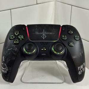 May include: Black video game controller with green and red accents. The controller has the text "Bloodborne" and "Fear the Old Blood". The controller is on a clear plastic stand.