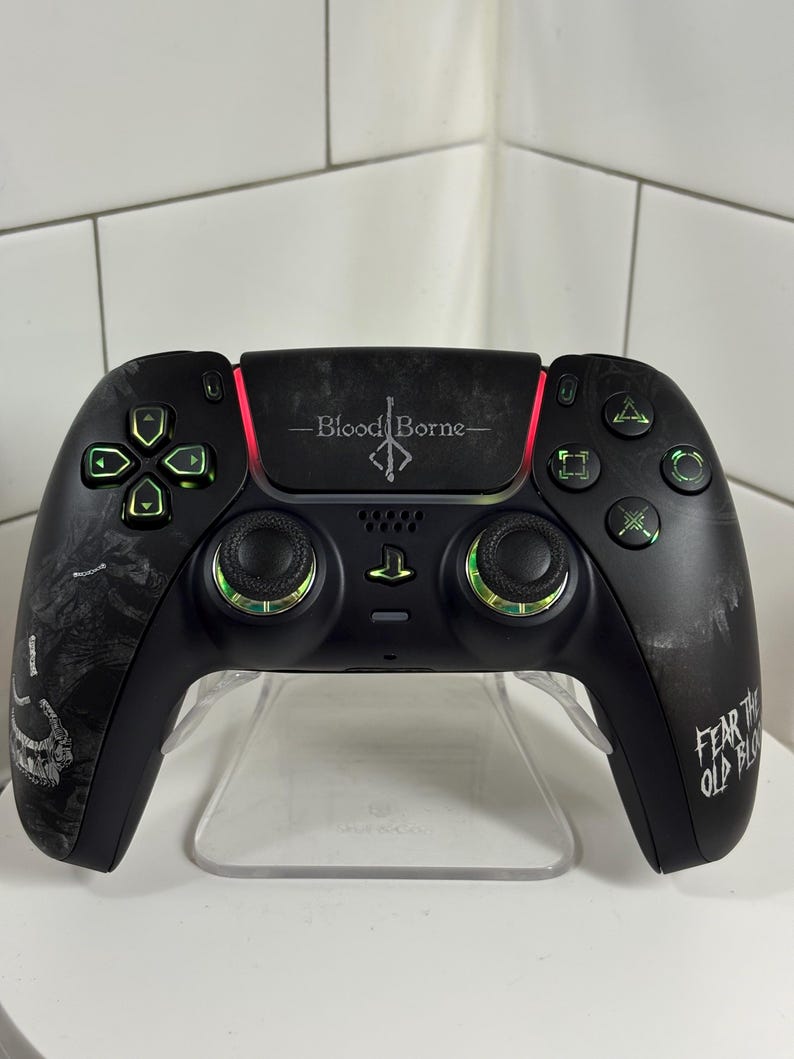 May include: Black video game controller with green and red accents. The controller has the text "Bloodborne" and "Fear the Old Blood". The controller is on a clear plastic stand.