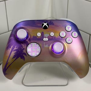 May include: Custom Xbox controller with a sunset-themed design. The controller features a gradient of purple, pink, and gold, with a silhouette of palm trees. The buttons are white and the thumbsticks have a purple glow.