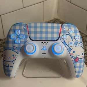 Puede incluir: Un mando de PlayStation 5 blanco y azul con un patrón de cuadros vichy azul claro. El mando presenta un diseño de personaje de dibujos animados y botones azules brillantes. El mando está sobre un soporte transparente.