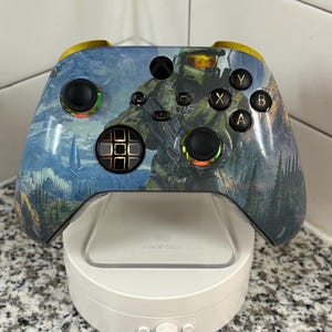 May include: A wireless video game controller featuring a graphic print of a green armored soldier in a landscape. The controller has black joysticks, a black directional pad, and buttons labeled A, B, X, and Y. It rests on a white circular stand.