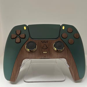 Puede incluir: Un mando de PlayStation 5 de color verde oscuro y marrón con detalles dorados. El mando está en un soporte transparente.