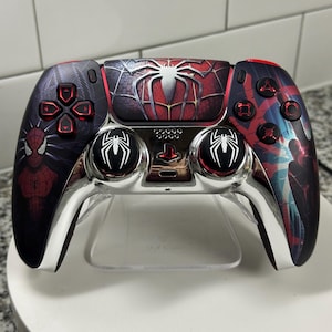May include: Customized PlayStation 5 controller featuring a Spider-Man design. The controller has a red, black, and silver color scheme with Spider-Man graphics. The buttons are red, and the joysticks have white spider emblems. The controller is displayed on a clear stand.
