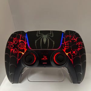 Puede incluir: Un mando de PlayStation 5 negro con un diseño de telaraña y un gráfico de araña en la parte superior. El mando está iluminado con luces rojas y azules.