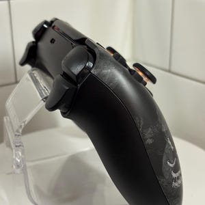 May include: Black gaming controller with a custom design on the side, featuring black buttons and orange accents. The controller is displayed on a clear acrylic stand, showcasing its ergonomic design and detailed features.