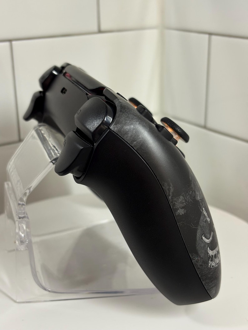 May include: Black gaming controller with a custom design on the side, featuring black buttons and orange accents. The controller is displayed on a clear acrylic stand, showcasing its ergonomic design and detailed features.