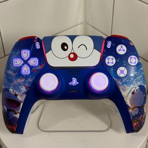 Op de afbeelding: Een blauw-rode PlayStation 5-controller met een cartoonkarakterontwerp. De controller heeft een blauwe achtergrond met een cartoonkarakter aan de voorkant. De knoppen zijn blauw en wit. De controller staat op een transparante standaard.