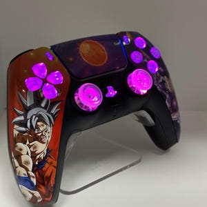 Puede incluir: Un mando de PlayStation 5 negro con una piel personalizada que presenta un personaje del anime Dragon Ball Z. El mando tiene botones de color púrpura brillante y un fondo púrpura.