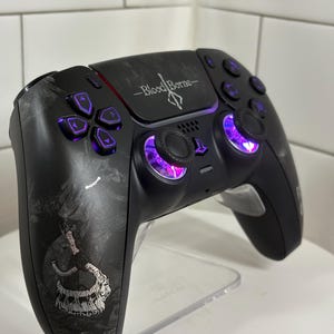 May include: Black video game controller with purple buttons and joysticks. The controller has a graphic design and the words "Bloodborne" are printed on the top. The controller is on a clear stand.