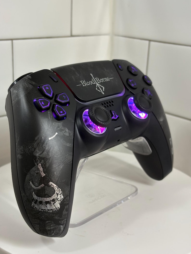 May include: Black video game controller with purple buttons and joysticks. The controller has a graphic design and the words "Bloodborne" are printed on the top. The controller is on a clear stand.