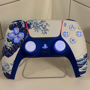 May include: A white and blue video game controller with a wave pattern design. The controller has blue buttons and accents, with a PlayStation logo in the center. The controller is on a clear stand.