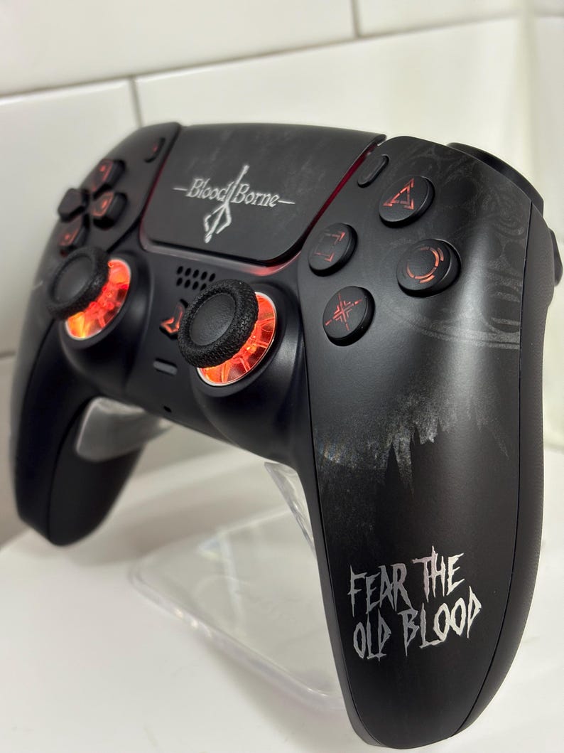 May include: Black gaming controller with the words "Bloodborne" and "Fear the Old Blood" in white. The controller has red accents around the joysticks and buttons. The controller is designed for video games.