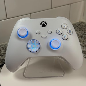 Peut inclure: Une manette Xbox blanche avec des accents bleus illuminés sur les joysticks et les boutons. La manette présente le logo Xbox et une disposition standard des boutons. Elle est présentée sur un support transparent.