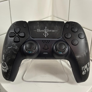 May include: Black video game controller with the text "Bloodborne" and "Fear the Old Blood." The controller has a unique design with detailed graphics and a clear stand.
