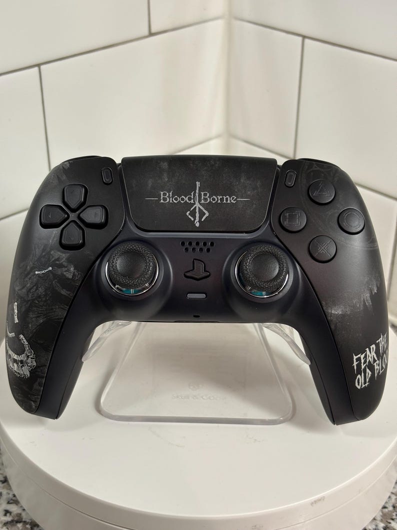 May include: Black video game controller with the text "Bloodborne" and "Fear the Old Blood." The controller has a unique design with detailed graphics and a clear stand.