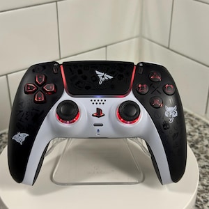 May include: A black and white video game controller with red accents. The controller features a white center section with black buttons and directional pads. The black sections have various graphic designs. The controller is on a clear stand.