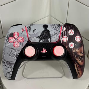 Può includere: Controller PlayStation 5 personalizzato con base nera e dettagli bianchi e marroni. Il controller presenta opere d'arte della serie anime "Attack on Titan", con pulsanti illuminati di rosso e un supporto trasparente.