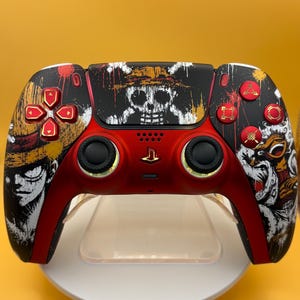 Può includere: Un controller per videogiochi personalizzato con una combinazione di colori rosso e nero e grafiche di One Piece. Il touchpad centrale mostra un logo bianco con teschio e ossa incrociate, mentre le impugnature presentano illustrazioni.