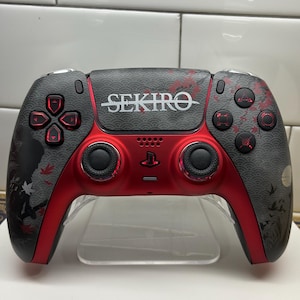 Può includere: Un controller PlayStation 5 personalizzato, nero e rosso. Il controller presenta la scritta "SEKIRO" in bianco, pulsanti rossi e un design di foglie d'acero giapponesi. Su un supporto trasparente.
