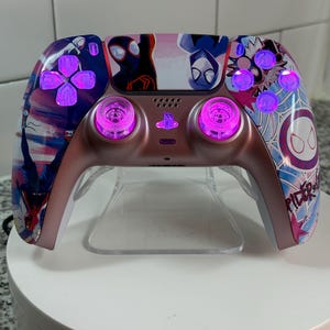 May include: A custom-designed video game controller featuring a Spider-Man theme. The controller has a rose gold finish with a vibrant graphic design of Spider-Man characters. The buttons and joysticks glow with a bright pink light, enhancing the visual appeal.