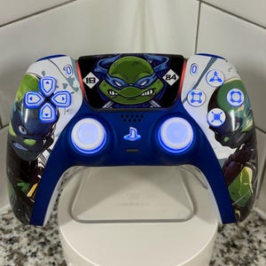 May include: A custom blue and white PlayStation 5 controller featuring Teenage Mutant Ninja Turtles artwork. The controller has blue LED lights and a charging stand. The design includes images of the Ninja Turtles and their iconic masks.