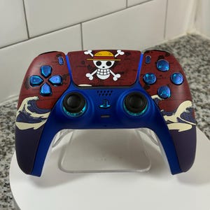 May include: A custom video game controller with a red and blue color scheme. The controller features a skull and crossbones design, blue buttons, and a wave pattern. The controller is displayed on a clear stand.