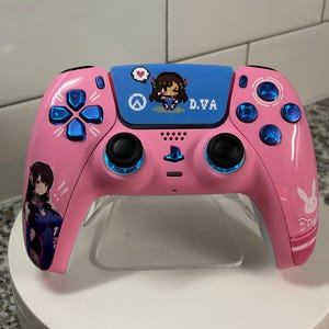 May include: Pink video game controller with blue accents and black joysticks. The controller features a pixelated character design and the text "D.VA". Additional graphics include a heart, a peace symbol, and a bunny.