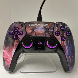 Könnte beinhalten: Ein schwarzer PlayStation 5-Controller mit einem Tekken 8-Design. Der Controller hat violette Lichter um die Tasten und die Analogsticks. Der Controller steht auf einem durchsichtigen Ständer.