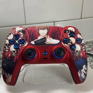 May include: Red and black video game controller with a Death Note theme. The controller features a design with skulls, roses, and anime characters. The buttons are blue and the controller is on a clear stand.
