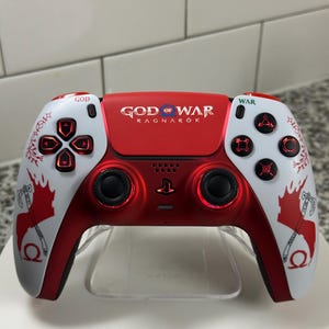 May include: A custom red and white PlayStation 5 controller. The controller features the words "GOD OF WAR RAGNAROK" across the top. It has red buttons and accents, with white side panels featuring axe and character designs.